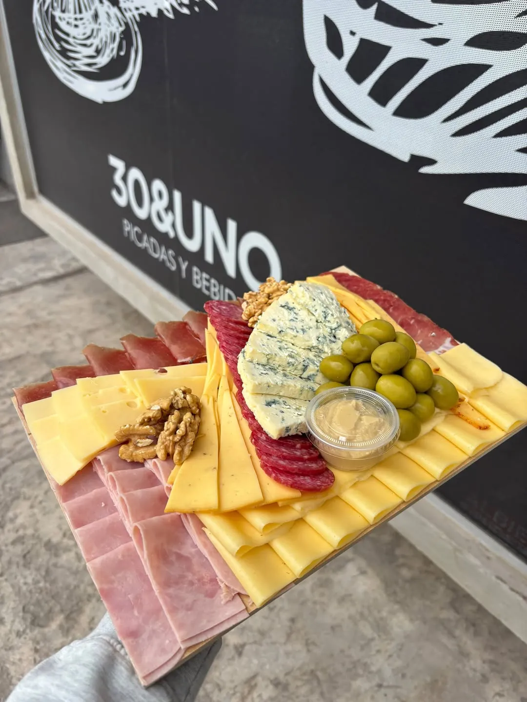 Tabla de picada con jamón cocido, quesos en abanico, salame, roquefort, nueces, aceitunas verdes y dip, sostenida frente al cartel negro de la fachada de 30&UNO