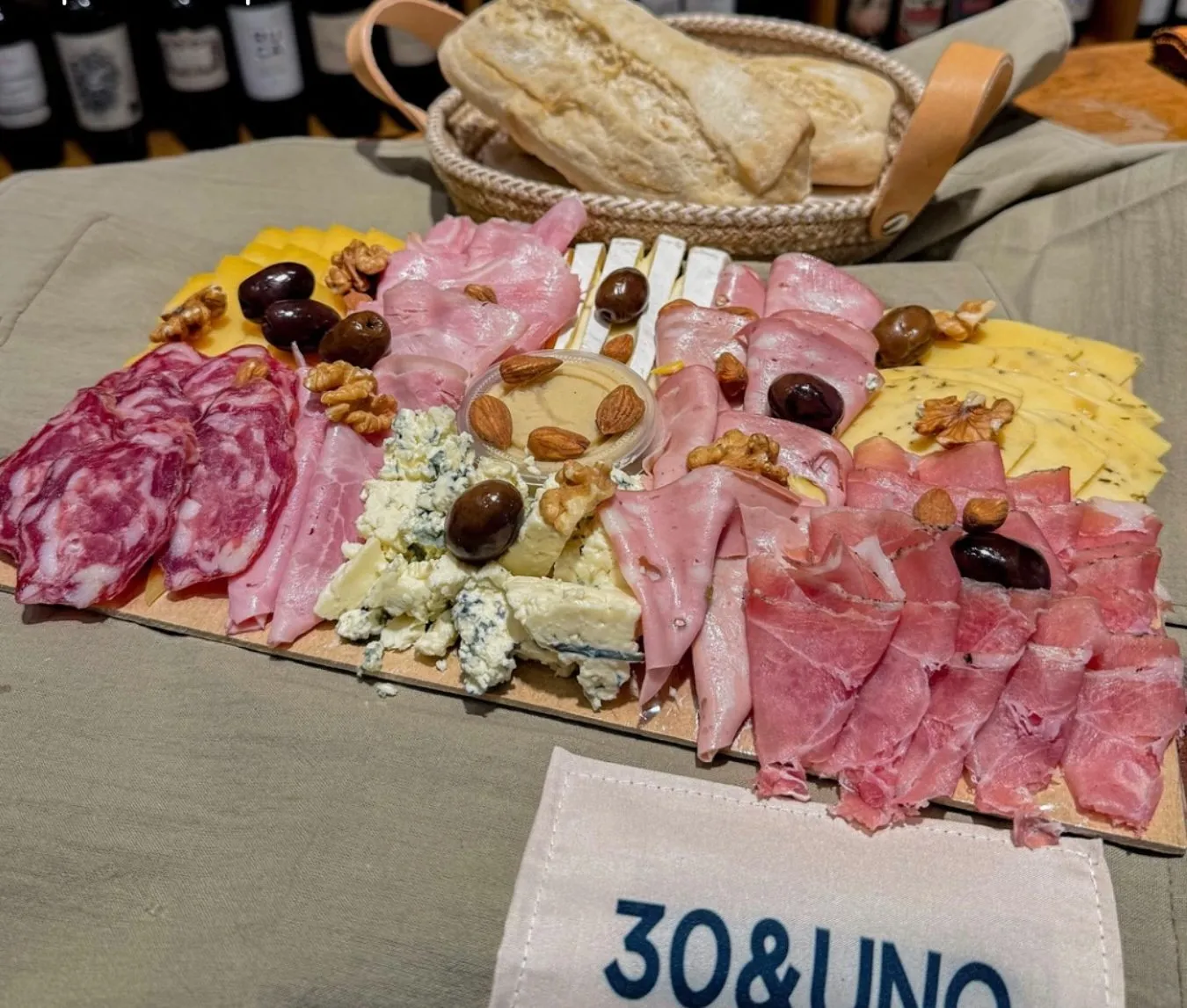 Picada Premium para 4 personas sobre lienzo verde oliva, con canasta de pan y estantes de vinos de fondo