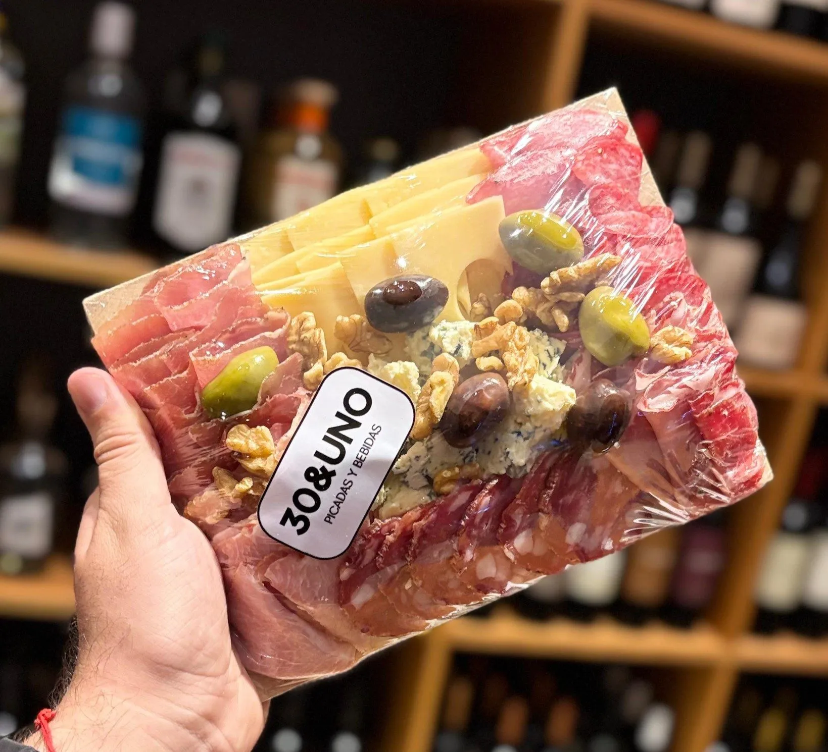 Mano sosteniendo la Picada Mediterránea para 2 envuelta en film con el sticker de 30&UNO, con la estantería de vinos de fondo