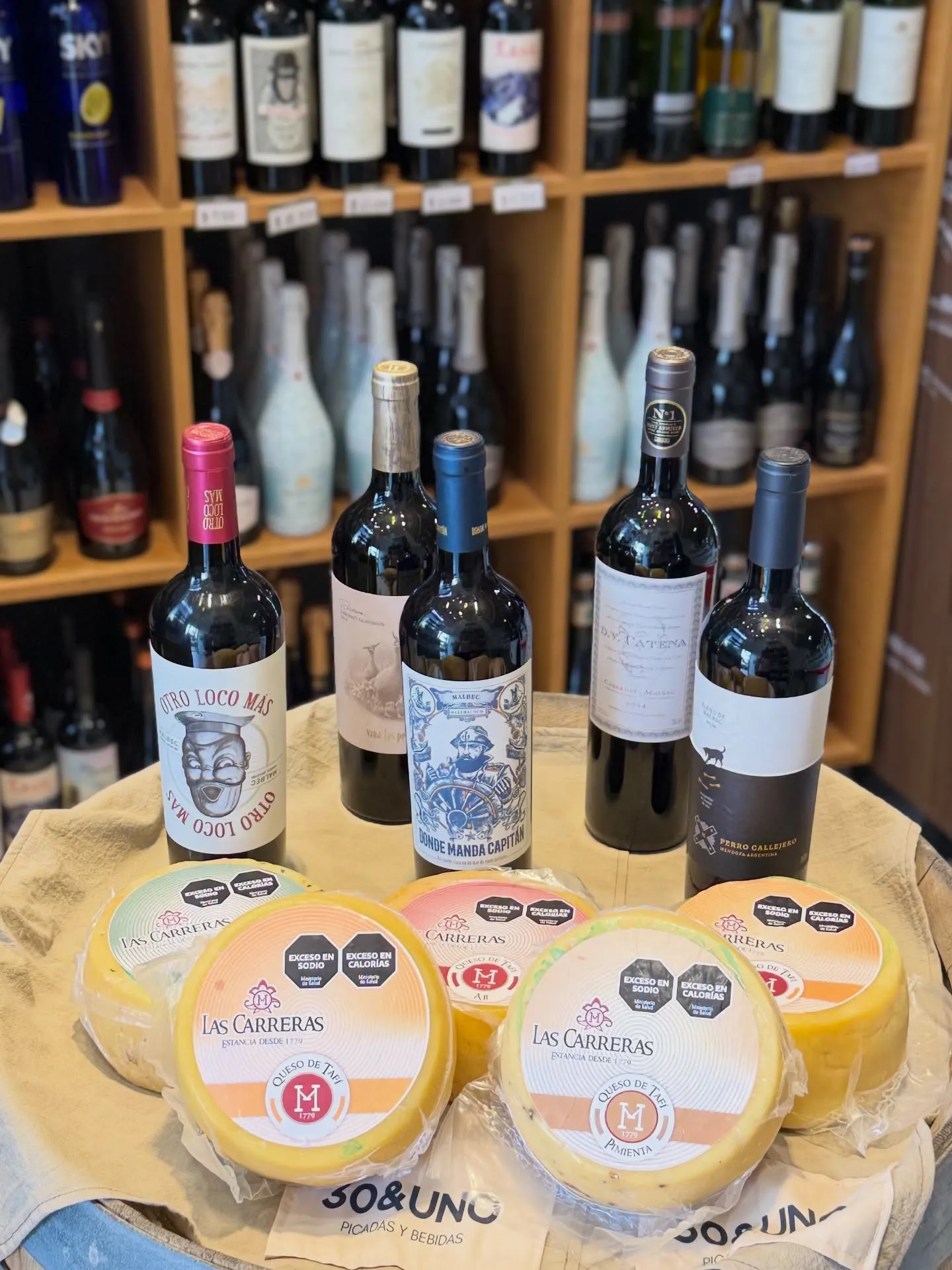 Cinco botellas de vino tinto (Otro Loco Más, Las Perdices, Donde Manda Capitán, D.V. Catena y Perro Callejero) junto a hormas de Queso de Tafí Las Carreras sobre una bolsa de tela con el logo de 30&UNO