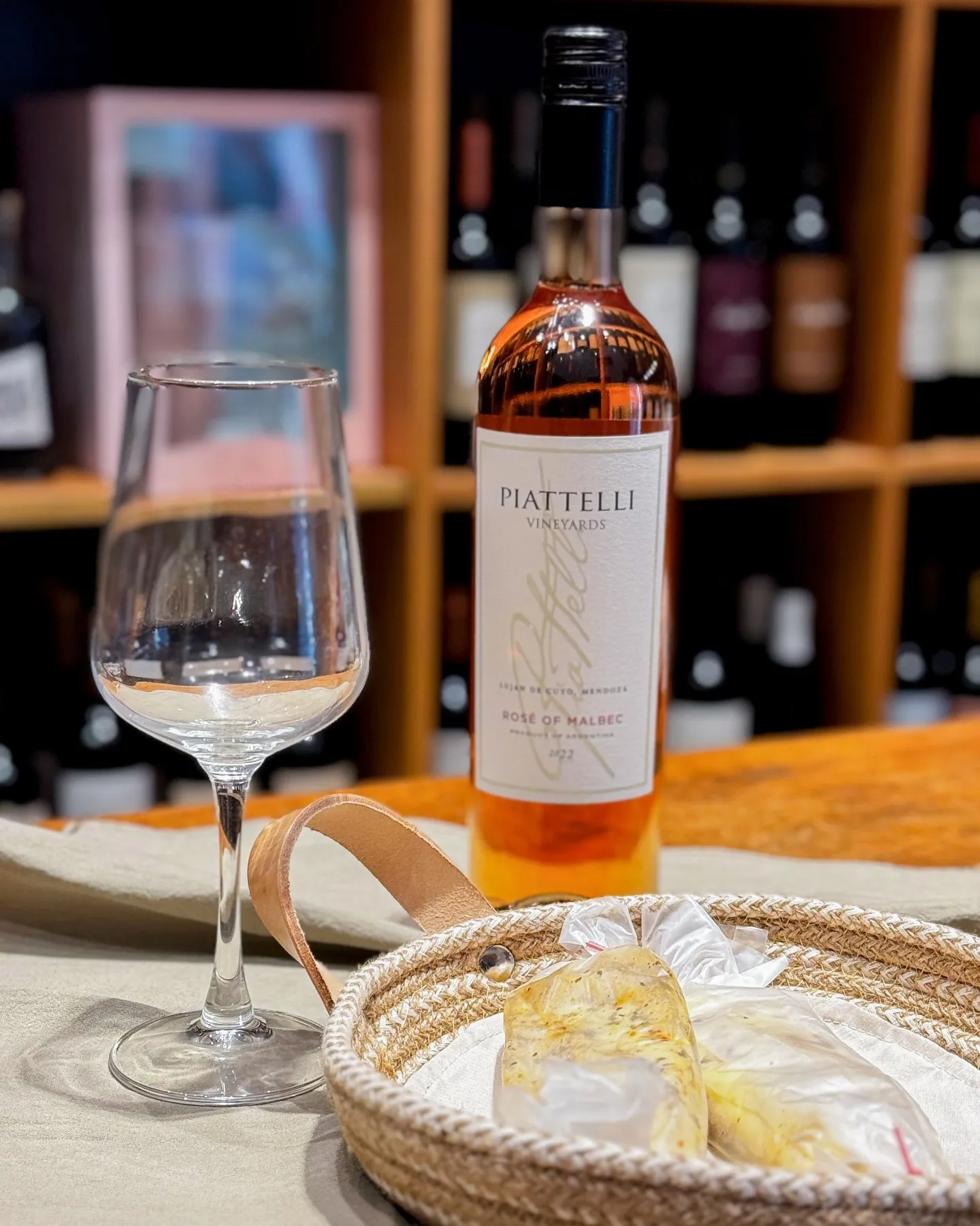 Botella de Piattelli Rosé of Malbec con una copa vacía y una canasta de mimbre con quesillo, estantes de vinos desenfocados de fondo