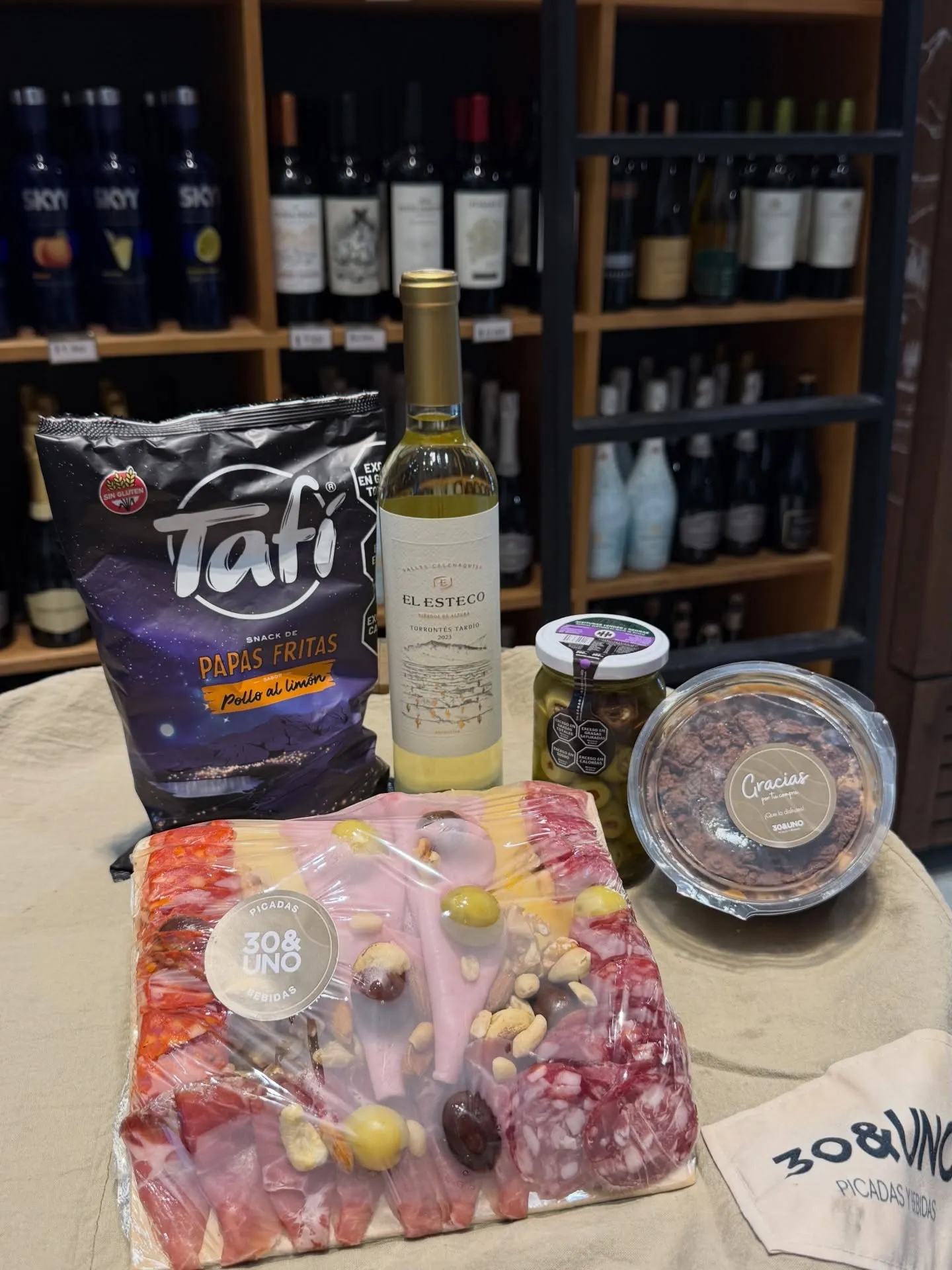 Box de regalo de 30&UNO: picada envuelta con sticker, botella de El Esteco Torrontés Tardío, frasco de aceitunas, papas Tafí y postre en pote con tarjeta, sobre lienzo beige con bolsa de tela